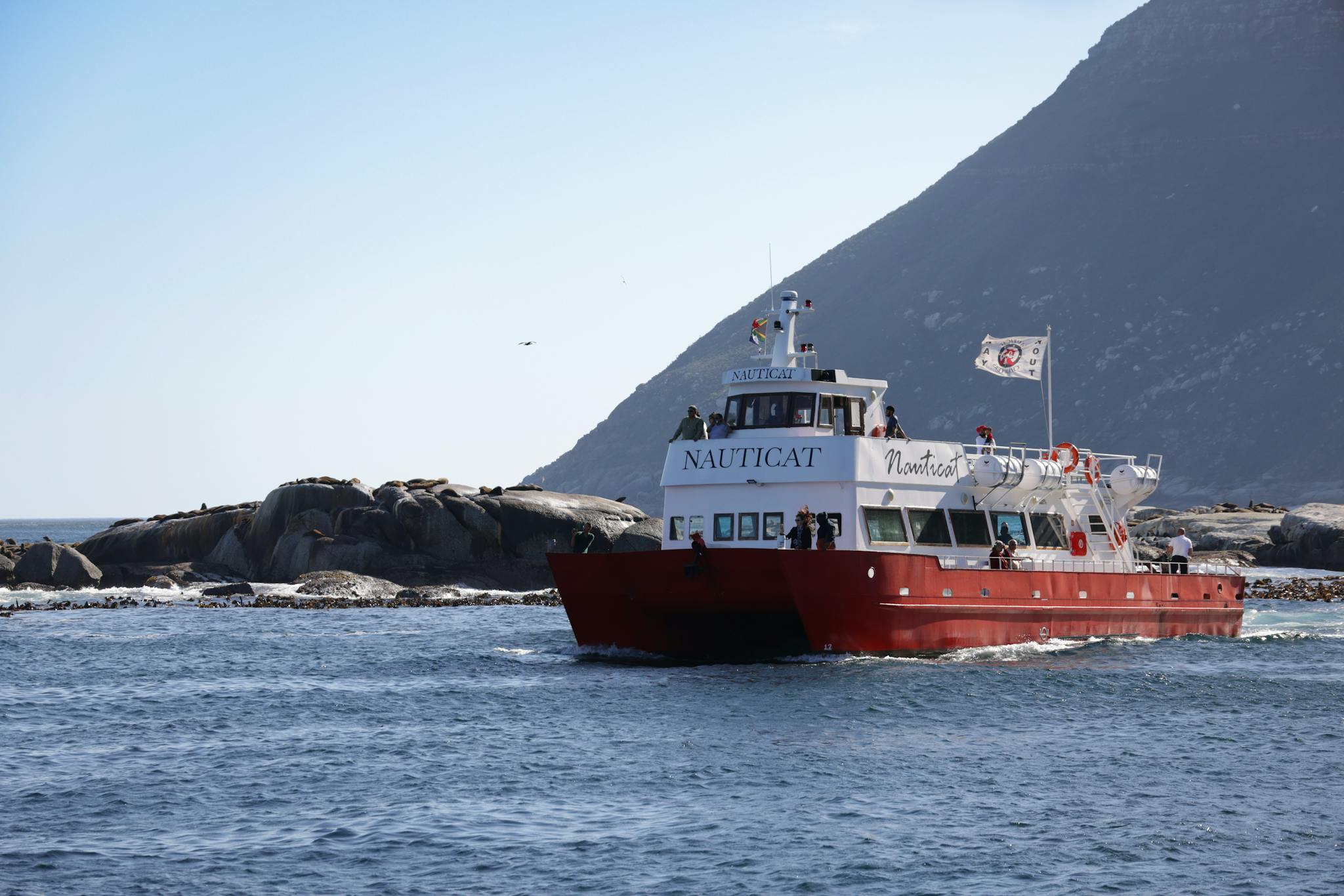 Red boat navigates near rocky shores with a mountainous backdrop in clear daylight.